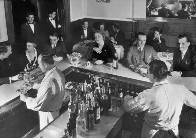 "Patrons & bartenders enjoying the ambiance of a gold & Victorian green bar while a Meyer Davis Orchestra (not seen) & a young lady sings in French & English at this popular unident. speakeasy, a haven for drinkers during prohibition."